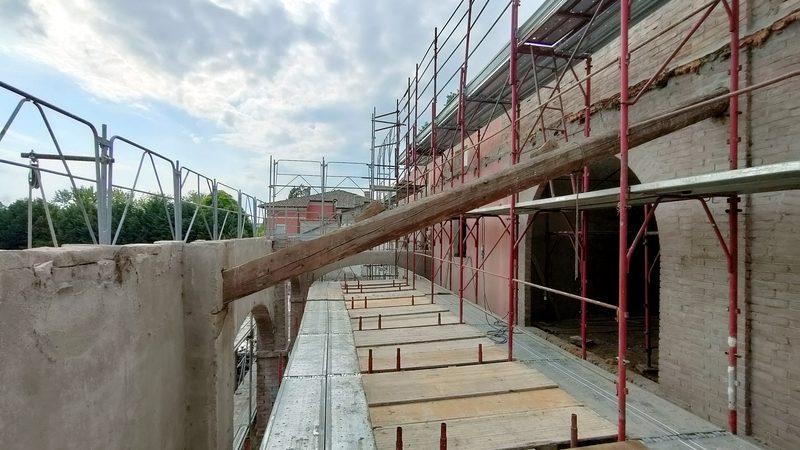 Edificio storico in muratura di mattoni con archi, in corso di recupero, con ponteggi lungo la facciata e un'antica trave in legno.
